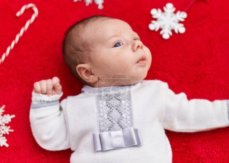 Photo for Adorable caucasian baby lying on floor with christmas decor over isolated red background - Royalty Free Image
