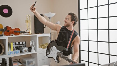 Téléchargez les photos : Jeune homme hispanique musicien jouant de la guitare électrique faire selfie par smartphone au studio de musique - en image libre de droit