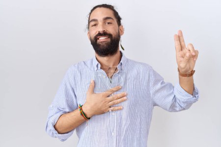 Foto de Hombre hispano con barba vistiendo camisa casual sonriendo jurando con la mano en el pecho y los dedos hacia arriba, haciendo una promesa de lealtad juramento - Imagen libre de derechos