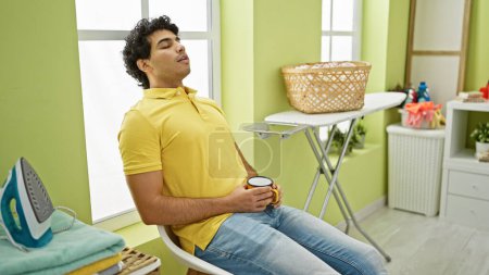 Téléchargez les photos : Jeune homme latin en attente de machine à laver tenant café dormant à la buanderie - en image libre de droit