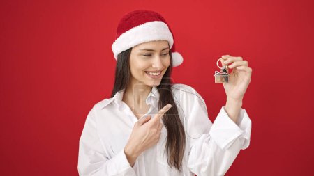 Téléchargez les photos : Jeune belle femme hispanique portant un chapeau de Noël pointant vers la clé de la nouvelle maison sur fond rouge isolé - en image libre de droit