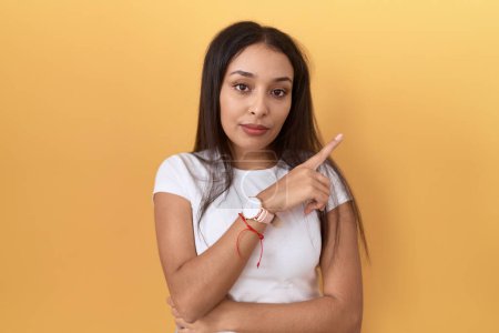 Photo for Young arab woman wearing casual white t shirt over yellow background pointing with hand finger to the side showing advertisement, serious and calm face - Royalty Free Image