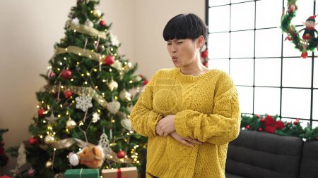 Photo for Young chinese woman suffering for stomachache standing by christmas tree at home - Royalty Free Image
