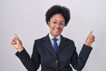 Téléchargez les photos : Belle femme africaine aux cheveux bouclés portant une veste d'affaires et des lunettes souriantes et confiantes pointant des doigts vers différentes directions. espace de copie pour la publicité - en image libre de droit