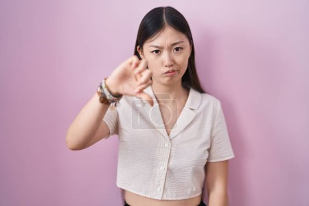 Foto de Mujer joven china de pie sobre fondo rosa mirando infeliz y enojado mostrando rechazo y negativo con los pulgares hacia abajo gesto. mala expresión. - Imagen libre de derechos
