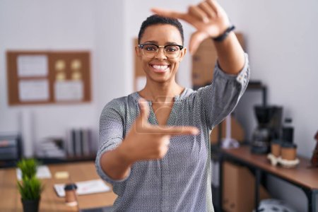 Foto de Mujer afroamericana trabajando en la oficina con gafas sonrientes haciendo montura con manos y dedos con cara feliz. concepto de creatividad y fotografía. - Imagen libre de derechos