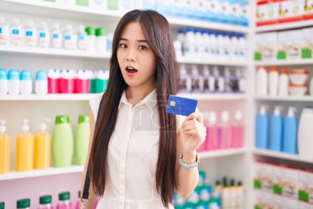 Foto de Joven mujer china de compras en farmacia con tarjeta de crédito asustada y sorprendida con la boca abierta para sorpresa, cara de incredulidad - Imagen libre de derechos