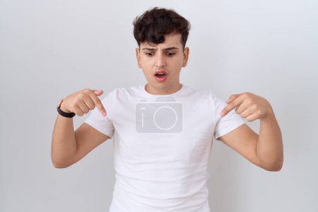 Téléchargez les photos : Jeune homme non binaire portant t-shirt blanc décontracté pointant vers le bas avec les doigts montrant la publicité, visage surpris et bouche ouverte - en image libre de droit