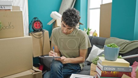 Photo for Young hispanic man writing on document sitting on sofa at new home - Royalty Free Image