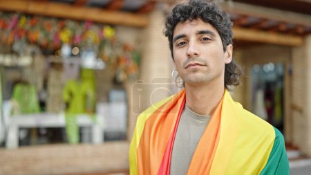Foto de Joven hombre hispano con bandera de arco iris con expresión relajada en la calle - Imagen libre de derechos