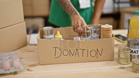 Téléchargez les photos : Tatoué, les mains gracieuses de l'homme hispanique volontaire, mettre des dons de nourriture en conserve dans une boîte en carton dans un centre de charité animé - le beau côté de travailler dans l'altruisme et l'unité - en image libre de droit