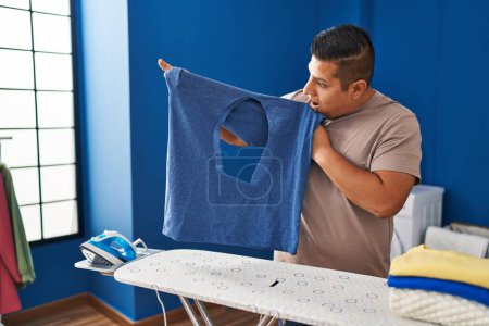 Foto de Joven latino sosteniendo camiseta con agujero quemado con expresión sorpresa en la lavandería - Imagen libre de derechos