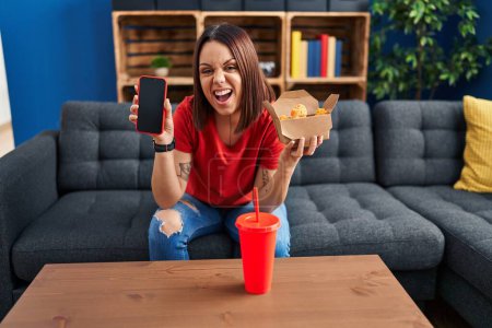 Foto de Mujer hispana joven comiendo comida rápida mostrando pantalla de teléfono inteligente enojado y loco gritando frustrado y furioso, gritando con ira. rabia y concepto agresivo. - Imagen libre de derechos