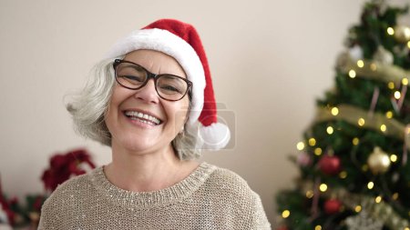 Téléchargez les photos : Femme d'âge moyen avec les cheveux gris souriant par arbre de Noël à la maison - en image libre de droit