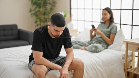 Photo for Beautiful couple disagreeing in their bedroom, sitting on the bed, expressing conflict while using a smartphone, a problem causing a serious expression - Royalty Free Image