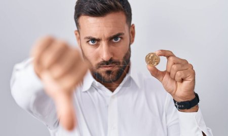 Foto de Hombre hispano guapo sosteniendo moneda criptomoneda litecoin con cara enojada, signo negativo que muestra aversión con los pulgares hacia abajo, concepto de rechazo - Imagen libre de derechos