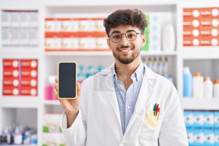 Téléchargez les photos : Homme arabe avec barbe travaillant à la pharmacie pharmacie montrant écran smartphone regardant positif et heureux debout et souriant avec un sourire confiant montrant les dents - en image libre de droit