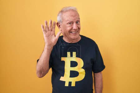 Téléchargez les photos : Homme âgé avec les cheveux gris portant Bitcoin t-shirt renonçant à dire bonjour heureux et souriant, geste de bienvenue convivial - en image libre de droit