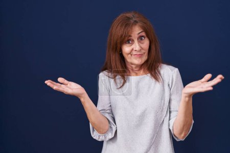 Photo for Middle age woman standing over blue background clueless and confused expression with arms and hands raised. doubt concept. - Royalty Free Image