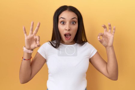 Photo for Young arab woman wearing casual white t shirt over yellow background looking surprised and shocked doing ok approval symbol with fingers. crazy expression - Royalty Free Image