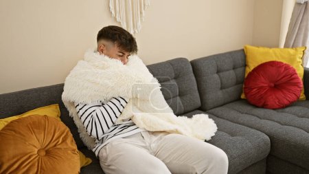Téléchargez les photos : Beau jeune homme hispanique gelant sur un canapé confortable à la maison, enveloppé dans une couverture chaude, combattant la grippe hivernale. portrait d'un homme séduisant à la recherche de confort dans la chaleur intérieure lorsque la température baisse. - en image libre de droit