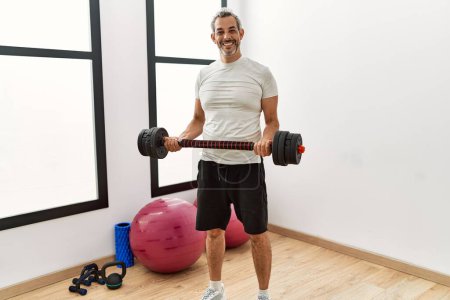 Téléchargez les photos : Homme aux cheveux gris d'âge moyen souriant confiant en utilisant la musculation au centre sportif - en image libre de droit