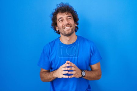 Foto de Hispanic young man standing over blue background hands together and fingers crossed smiling relaxed and cheerful. success and optimistic - Imagen libre de derechos