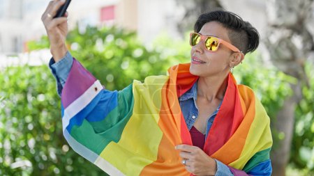 Foto de Joven hermosa mujer hispana hacer selfie por teléfono inteligente con bandera de arco iris en el parque - Imagen libre de derechos