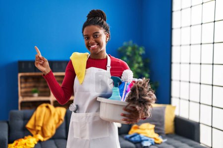 Photo for Young african american with braids cleaning the house holding cleaning products smiling happy pointing with hand and finger to the side - Royalty Free Image