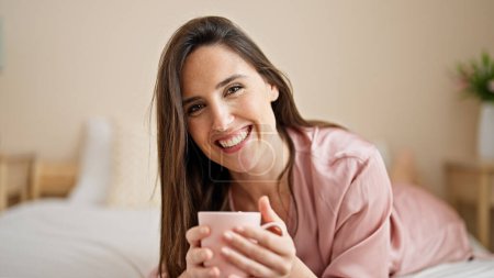 Téléchargez les photos : Jeune belle femme hispanique buvant une tasse de café allongée sur le lit parlant dans la chambre - en image libre de droit