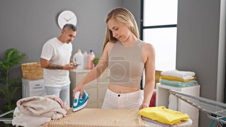 Photo for Man and woman couple ironing while boyfriend use smartphone at laundry room - Royalty Free Image