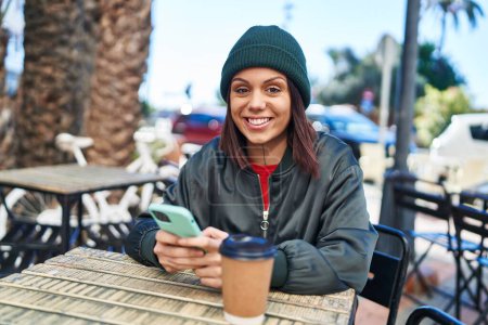 Foto de Young beautiful hispanic woman using smartphone drinking coffee at coffee shop terrace - Imagen libre de derechos