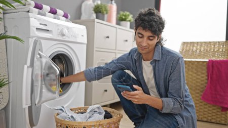 Photo for Young hispanic man using smartphone washing clothes at laundry room - Royalty Free Image