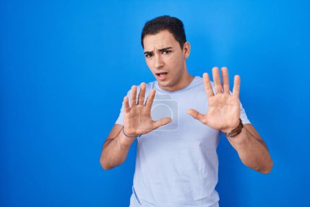 Photo for Young hispanic man standing over blue background afraid and terrified with fear expression stop gesture with hands, shouting in shock. panic concept. - Royalty Free Image
