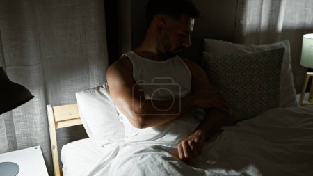 Téléchargez les photos : Jeune homme arabe assis sur le lit bras à gratter pour les démangeaisons à la chambre - en image libre de droit