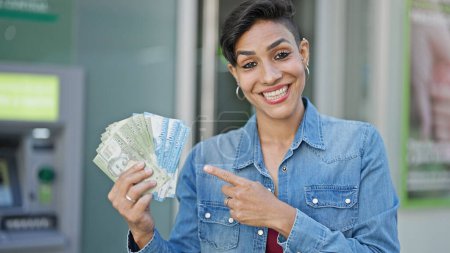 Foto de Joven hermosa mujer hispana sonriendo confiada señalando billetes chilenos en cajero de banco - Imagen libre de derechos