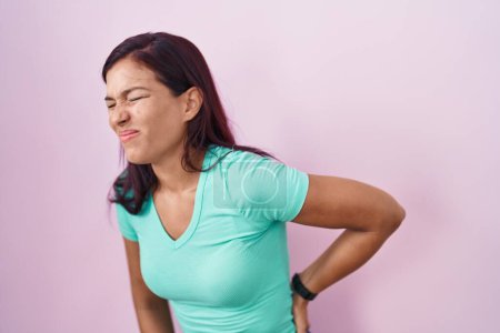 Téléchargez les photos : Jeune femme hispanique debout sur fond rose souffrant de maux de dos, toucher le dos avec la main, douleur musculaire - en image libre de droit