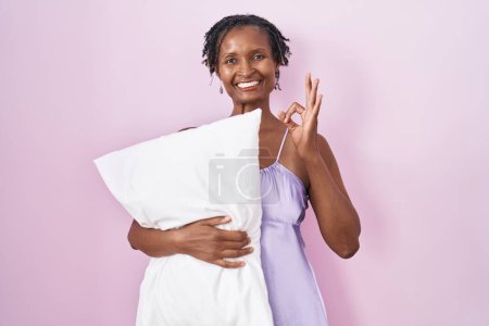 Photo for African woman with dreadlocks wearing pajama hugging pillow smiling positive doing ok sign with hand and fingers. successful expression. - Royalty Free Image