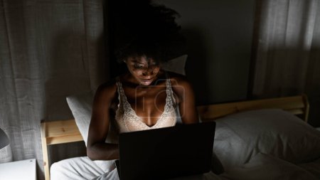 Foto de Mujer afroamericana usando lencería usando portátil sentado en la cama en el dormitorio - Imagen libre de derechos