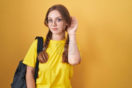 Photo for Young caucasian woman wearing student backpack over yellow background smiling with hand over ear listening an hearing to rumor or gossip. deafness concept. - Royalty Free Image