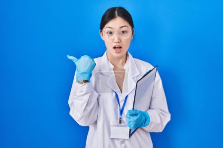 Foto de Mujer joven china que trabaja en el laboratorio científico sorprendido señalando con el dedo de la mano a un lado, boca abierta expresión asombrada. - Imagen libre de derechos