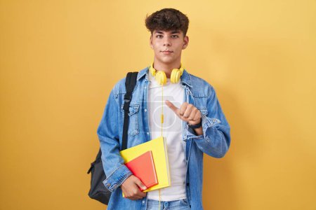 Foto de Adolescente hispana usando mochila de estudiante y sosteniendo libros apuntando hacia atrás con la mano y los pulgares hacia arriba, sonriendo confiada - Imagen libre de derechos