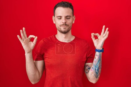 Téléchargez les photos : Jeune homme hispanique debout sur fond rouge détendu et souriant les yeux fermés faisant geste de méditation avec les doigts. concept de yoga. - en image libre de droit
