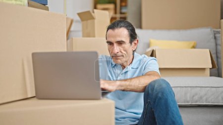 Téléchargez les photos : Homme d'âge moyen en utilisant un ordinateur portable assis sur le sol à la nouvelle maison - en image libre de droit