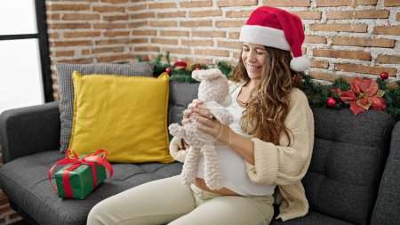 Photo for Young pregnant woman wearing christmas hat holding teddy bear at home - Royalty Free Image