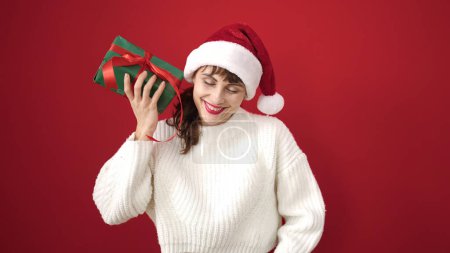Photo for Young caucasian woman smiling wearing christmas hat listening inside gift over isolated red background - Royalty Free Image