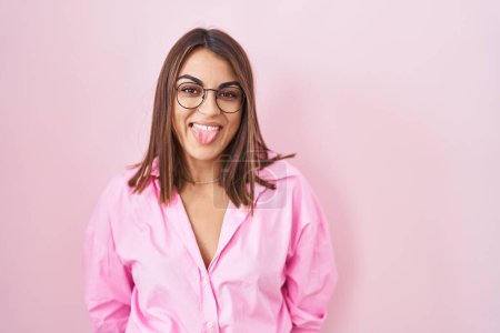 Téléchargez les photos : Jeune femme hispanique portant des lunettes debout sur fond rose langue collante heureux avec une expression drôle. concept d'émotion. - en image libre de droit