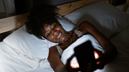 Téléchargez les photos : Femme afro-américaine portant de la lingerie en utilisant un smartphone couché sur le lit dans la chambre - en image libre de droit