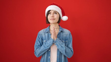 Foto de Joven mujer caucásica rezando usando sombrero de Navidad sobre fondo rojo aislado - Imagen libre de derechos