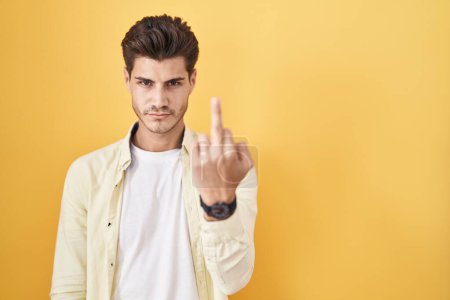 Foto de Young hispanic man standing over yellow background showing middle finger, impolite and rude fuck off expression - Imagen libre de derechos